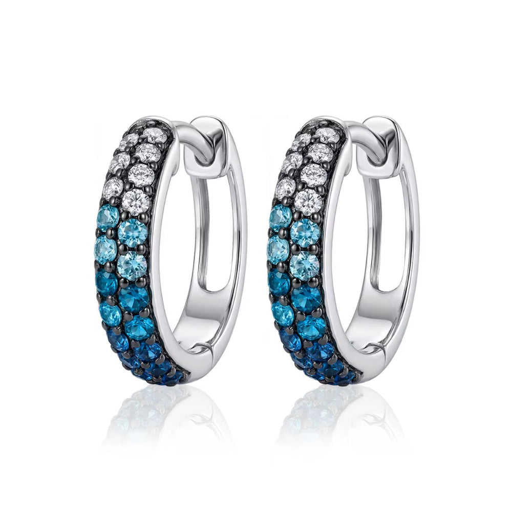 A pair of silver huggie hoop earrings featuring rows of clear cubic zirconia and blue topaz stones, displayed on a clean white background with a soft reflection.