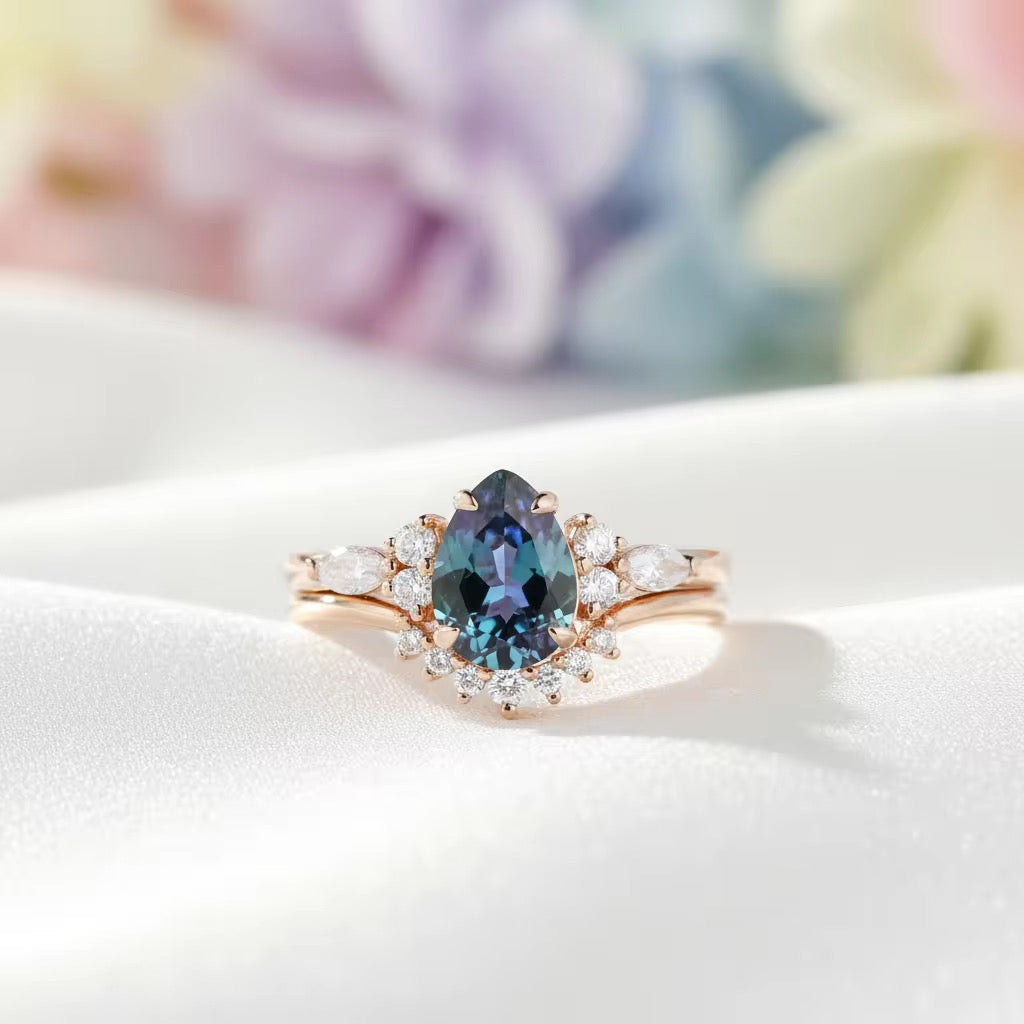A rose gold ring featuring a pear-cut blue gemstone surrounded by a halo of small clear stones, resting on a white satin surface with a soft, blurred pastel floral background.