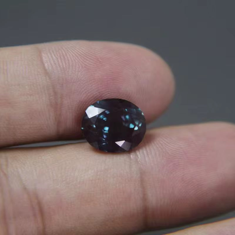 A round faceted Alexandrite gemstone resting on a person's fingers against a dark gray background.