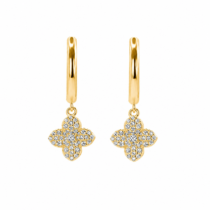A pair of gold huggie hoop earrings with dangling four-flower clover motifs paved with clear stones, shown on a solid white background.